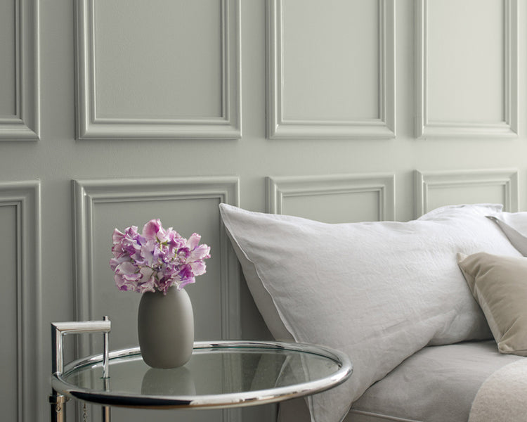 Gray wainscoting behind a bed with gray bedding and a glass side table featuring a small vase of purple flowers.